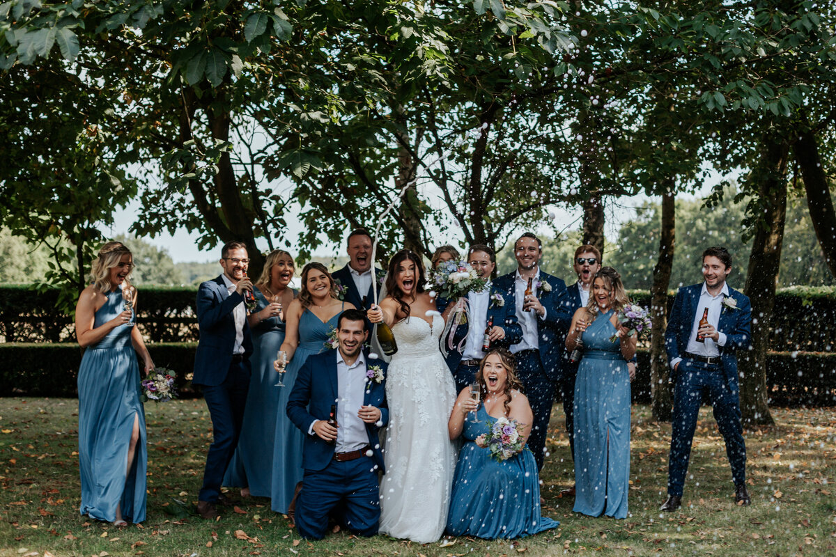 A wedding crew stood together cheering while the bride sprays champagne towards the camera