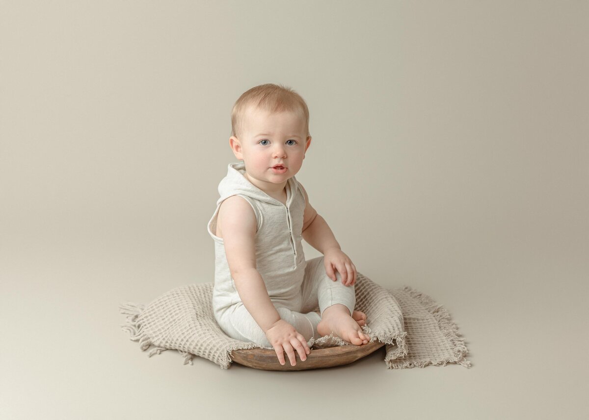 One year old boy dressed in oatmeal colored onesie holding rattle smiling