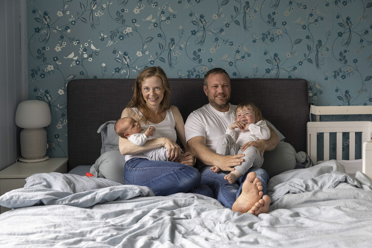 Livsstilsfoto familieportrett tatt på sengen. Mor og far holder hvert sitt barn og smiler mot kamera. Blå tapet bak sengen, grå headboard, lyseblått sengetøy.