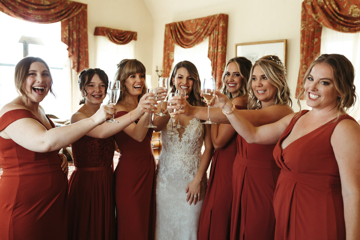 Bride drinks champagne with her bridesmaids in rust colored dresses.