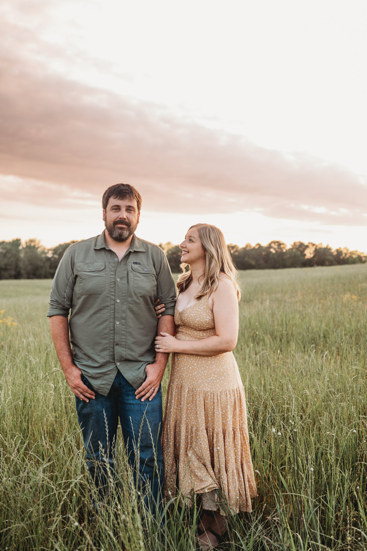 Wife gazes at husband during a family session in an open field with tall grass at sunset