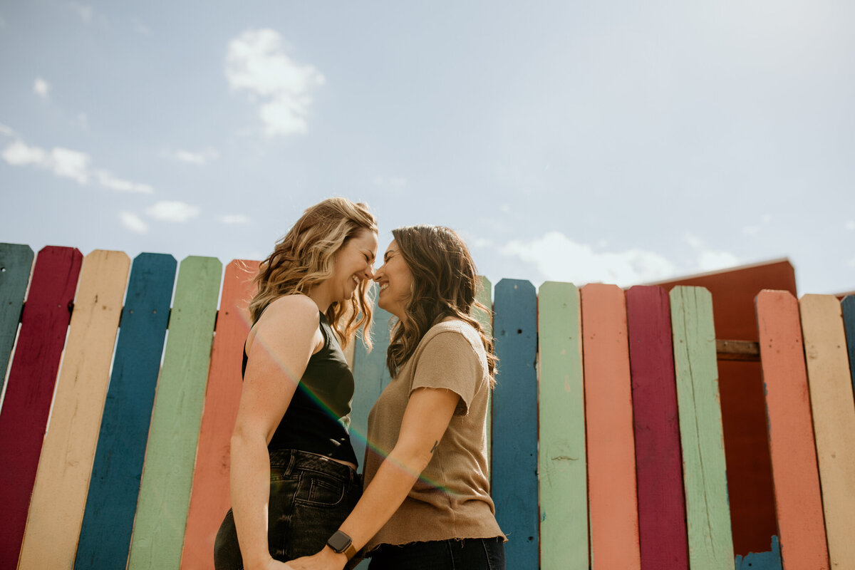 same sex couple about to kiss in front of a rainbow fence