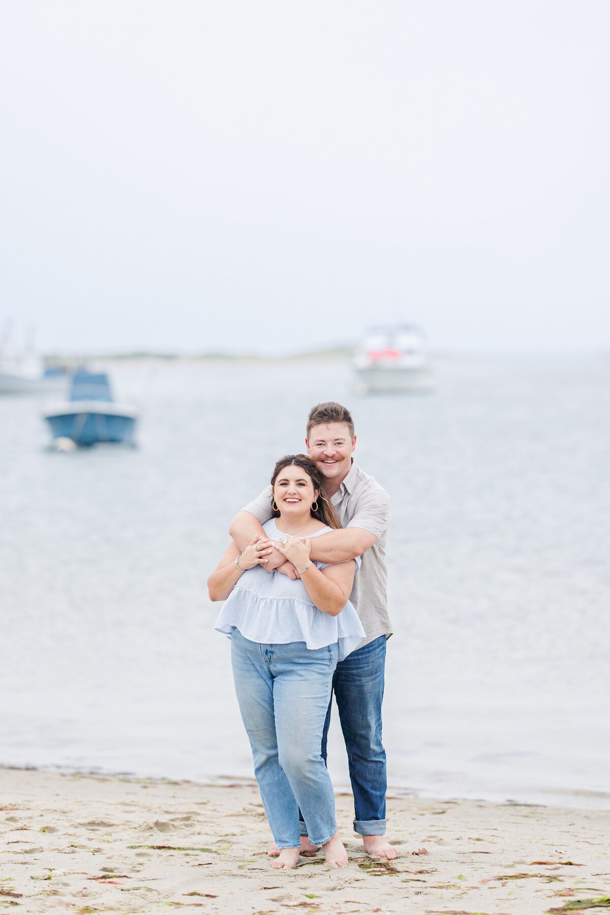 Man standing behind a woman on the beach, hugging her and kissing the top of her head, with anchored boats in the background at Chatham Bars Inn in Chatham, Massachusetts — representing the portfolio of a Cape Cod engagement photographer.