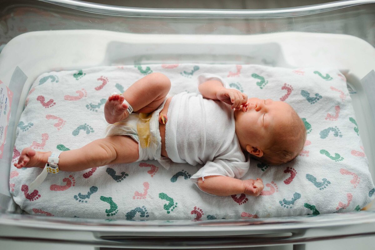 A serene image of a beautiful newborn baby peacefully sleeping in a transparent hospital cradle. The baby is dressed in a white shirt and lying on a soft white blanket, capturing a calm and tender moment.