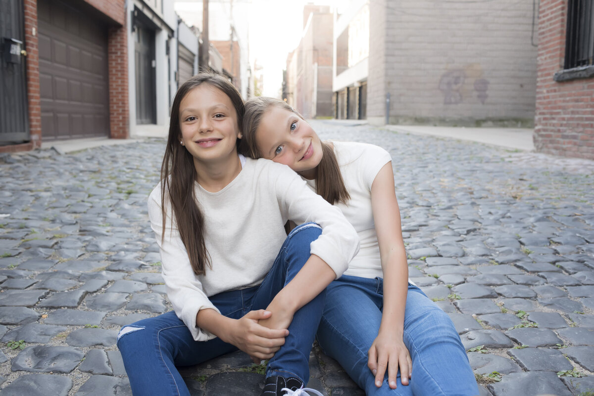 Two siblings sitting in an alley leaning into each other 