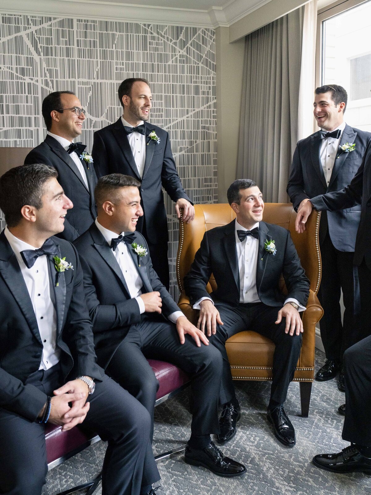 A group of seven men in tuxedos with bow ties and boutonnières gather in a room with modern patterned wallpaper. Some are seated on chairs, while others stand, all smiling and engaged in conversation. A large window brightens the scene.
