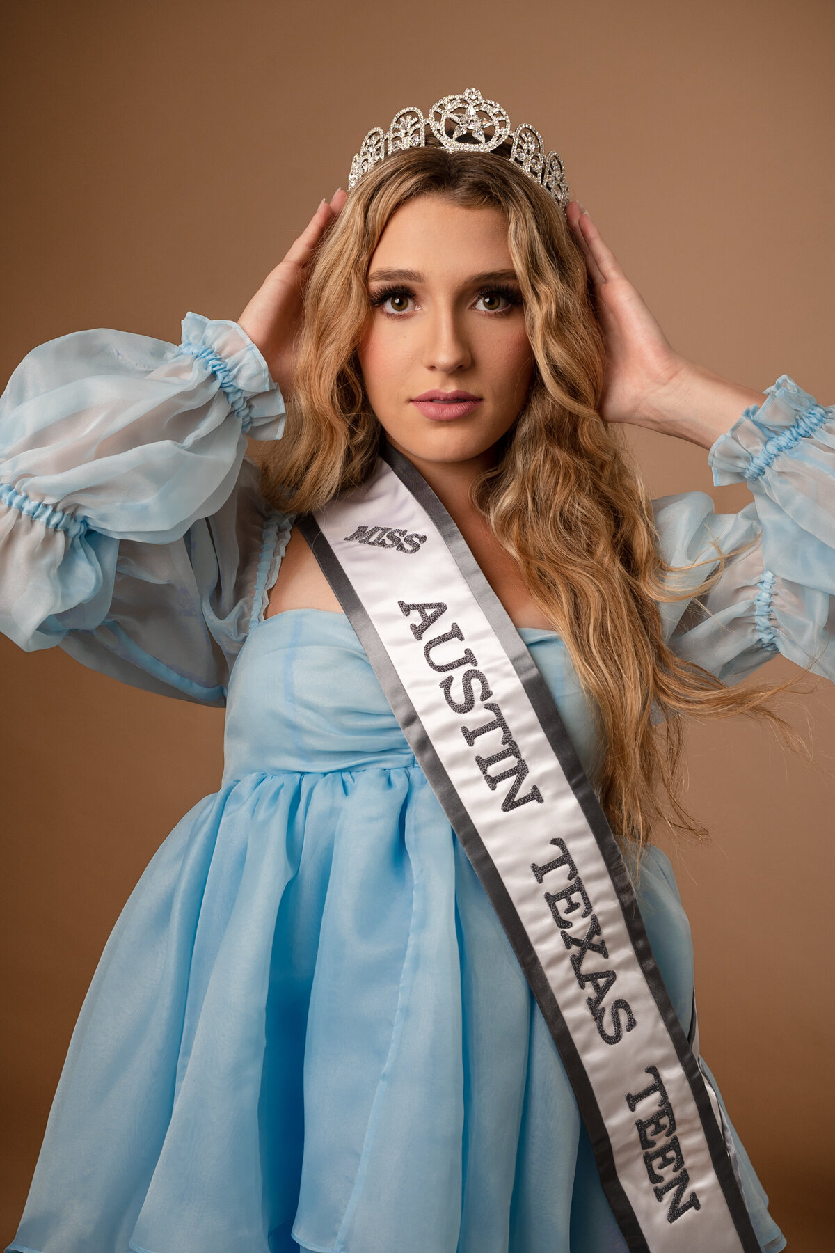 Miss Teen Austin poses wearing a blue soft dress and holding  crown with the two hands wearing a sash