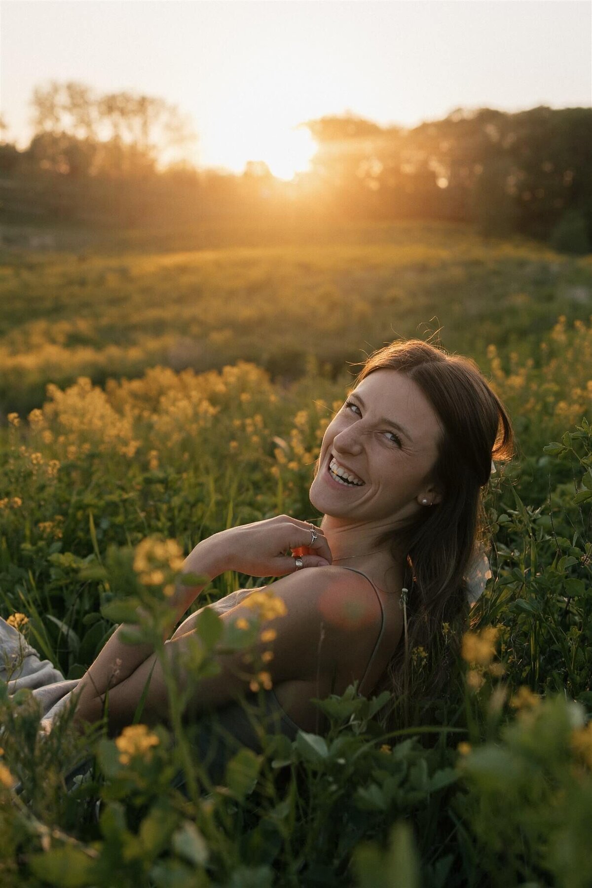 Wedding photographer looks over her shoulder and smiles as she sits in a sunlit meadow during golden hour