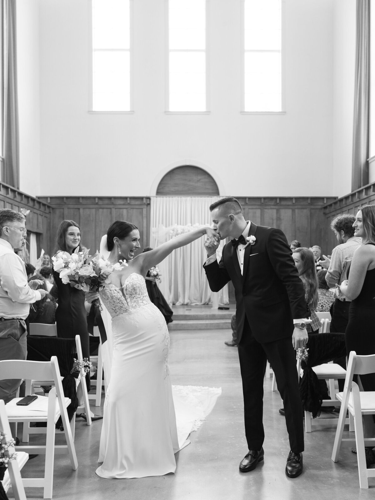 Bride raising bouquet as groom kisses her hand during joyful ceremony exit
