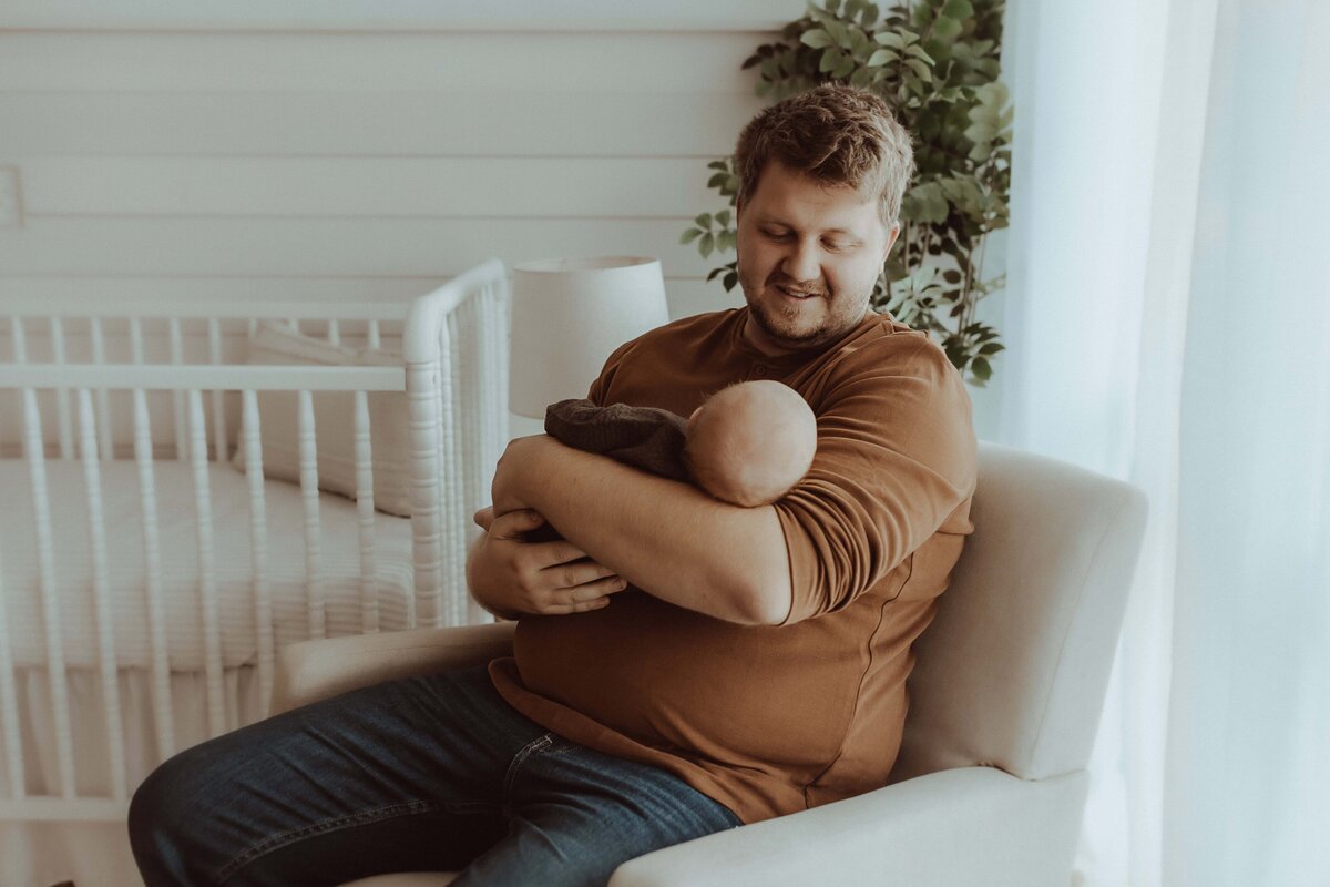 In a cozy Macon studio, a person sits in a chair cradling a baby, with a crib and a plant softly accentuating the background, capturing the essence of newborn photography.