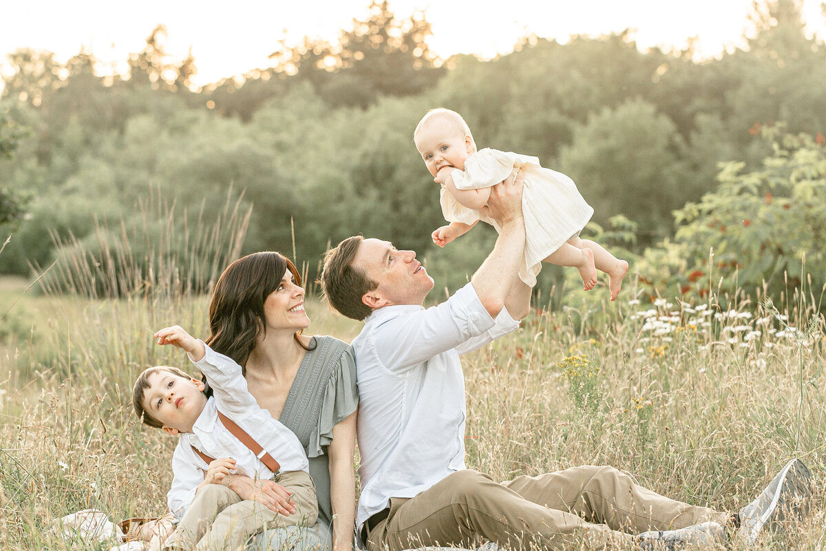 Family of four sitting down in a field of tall grasses all dressed in light, neutral tones and colors. Dad is holding up baby girl and mom is looking over at them. While brother is looking up at the sky. Sunset Summer Portland Family Photography Session.