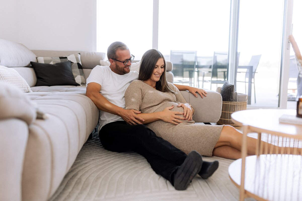 Man und Frau sitzen auf dem Boden vor ihrem Sofa, Frau hält ihren Babybauch