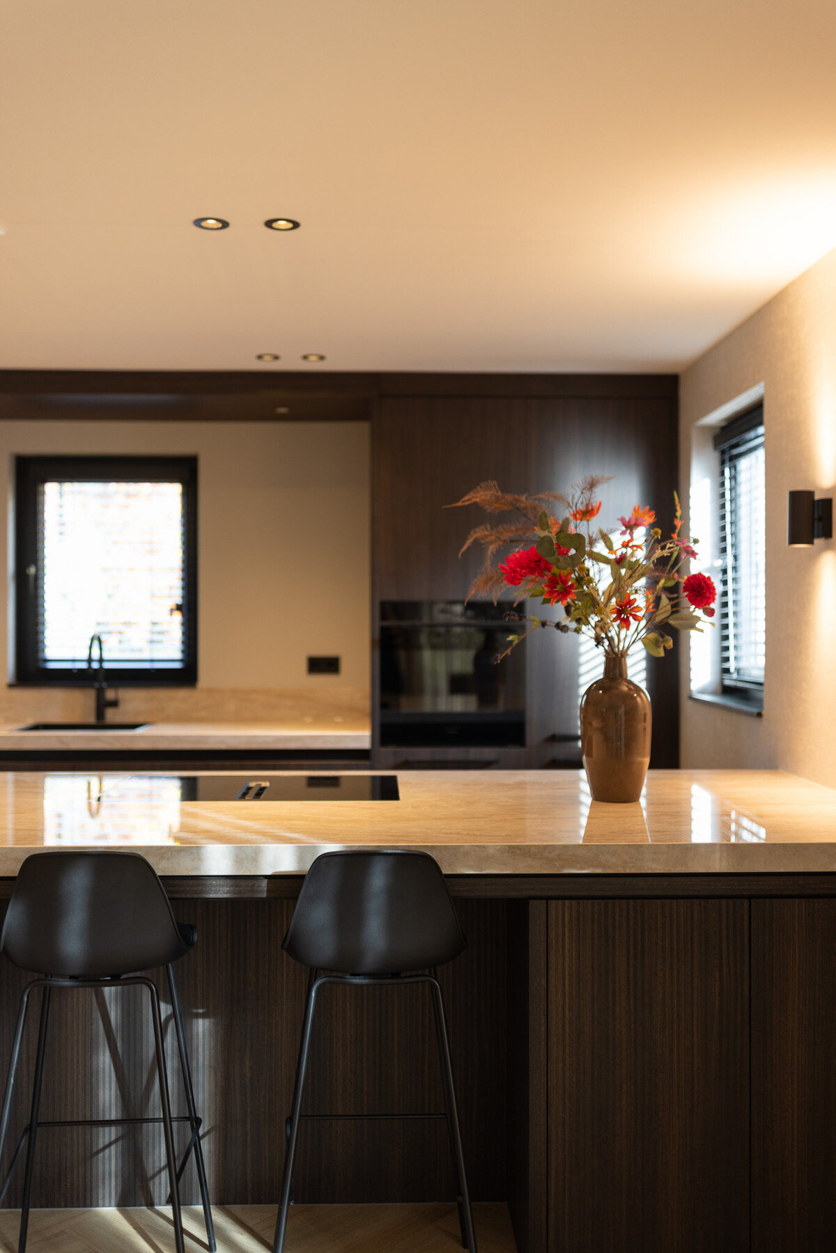 Warme keuken met donker houten fronten, een nis met verlichting en een stijlvol werkblad. De barstoelen en bloemen zorgen voor een huiselijke sfeer, terwijl het ontwerp strak blijft.