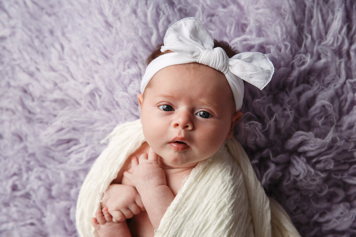 Beautiful newborn baby laying on a purple run and wearing a white headband and white blanket