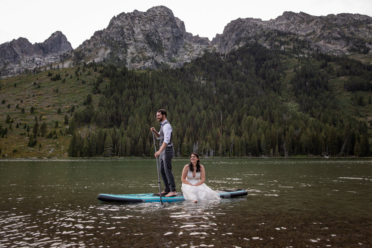 A bride sits on a paddle board while her groom paddles her around a lake in Wyoming.