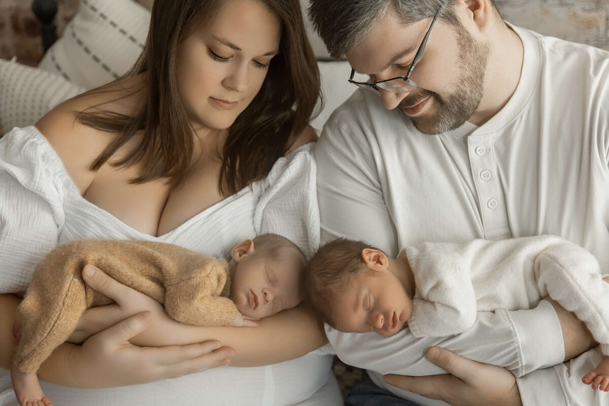 A mother and father sit close together, gazing at their sleeping newborn twins. The mother cradles one baby, while the father holds the other. They are dressed in soft, casual clothing, and the atmosphere feels warm and intimate, highlighting their joy and connection as a family.