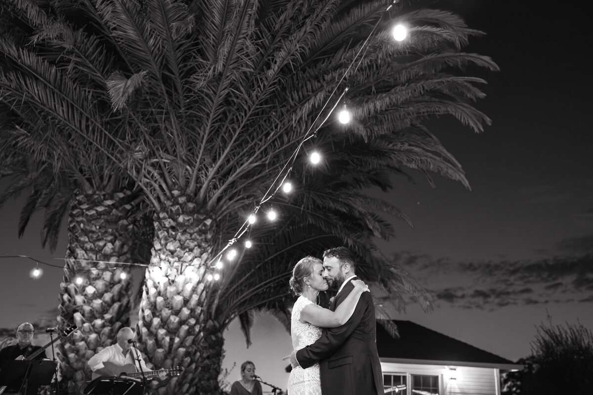 black and white photo of bride ang grooms first dance at sunset under string lights.  A big palm tree ais behind them, under the tree musicians play instruments in sonoma vineyard home