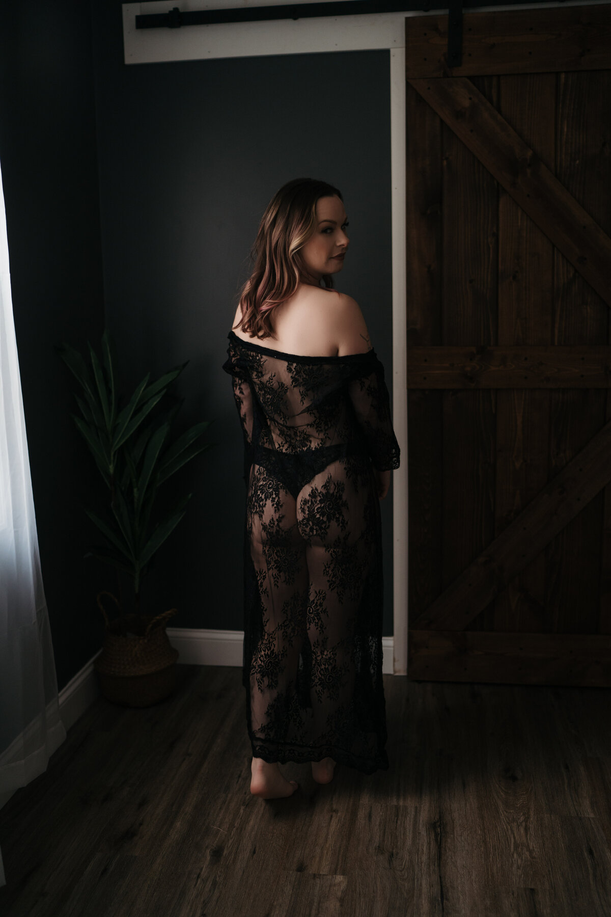 A woman in a black lace cover-up and black thong walks through a studio by a window looking over her shoulder