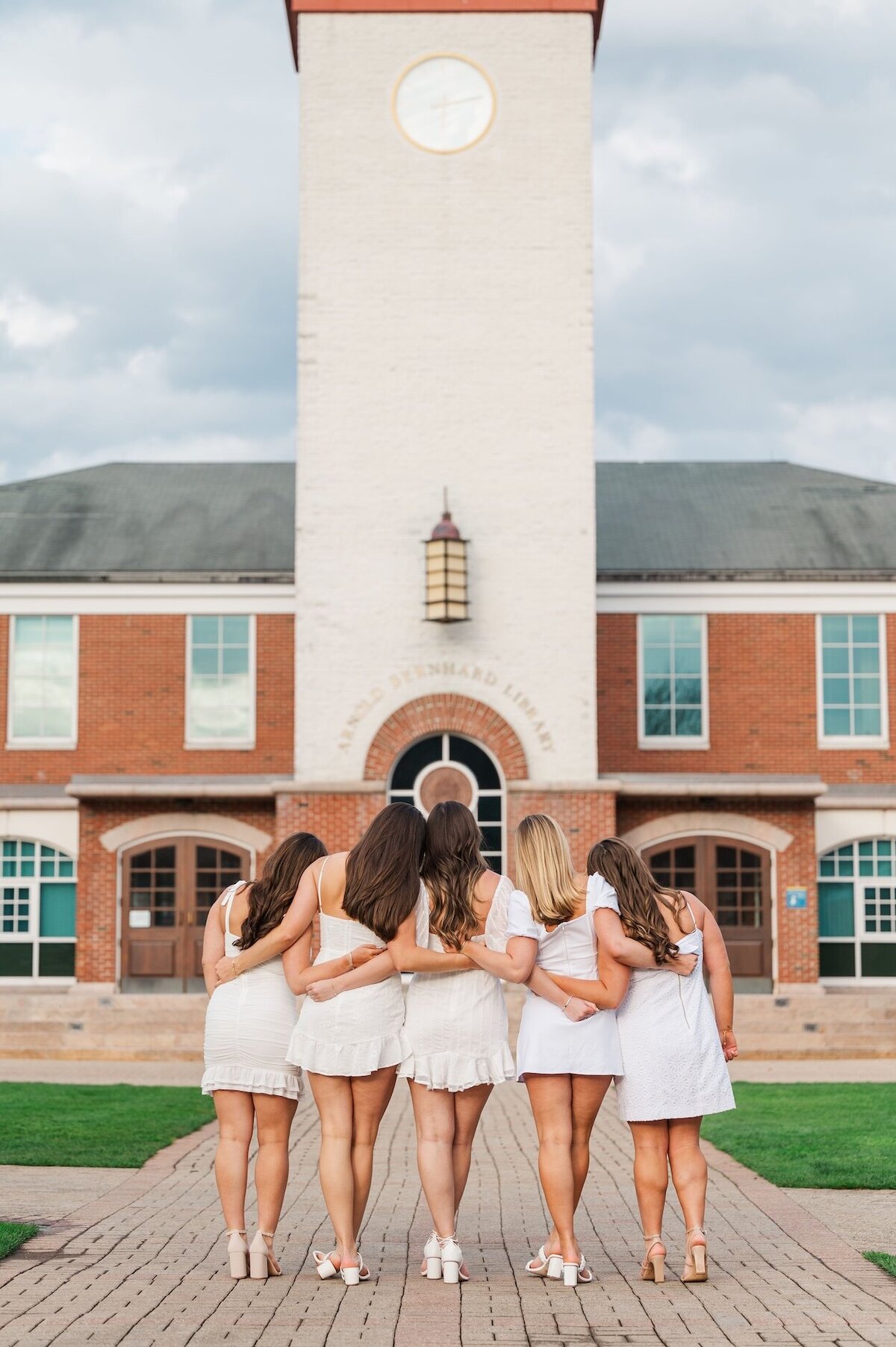 Five Quinnipiac graduates put their arms around one another as they look at Arnold Bernhard Library. 