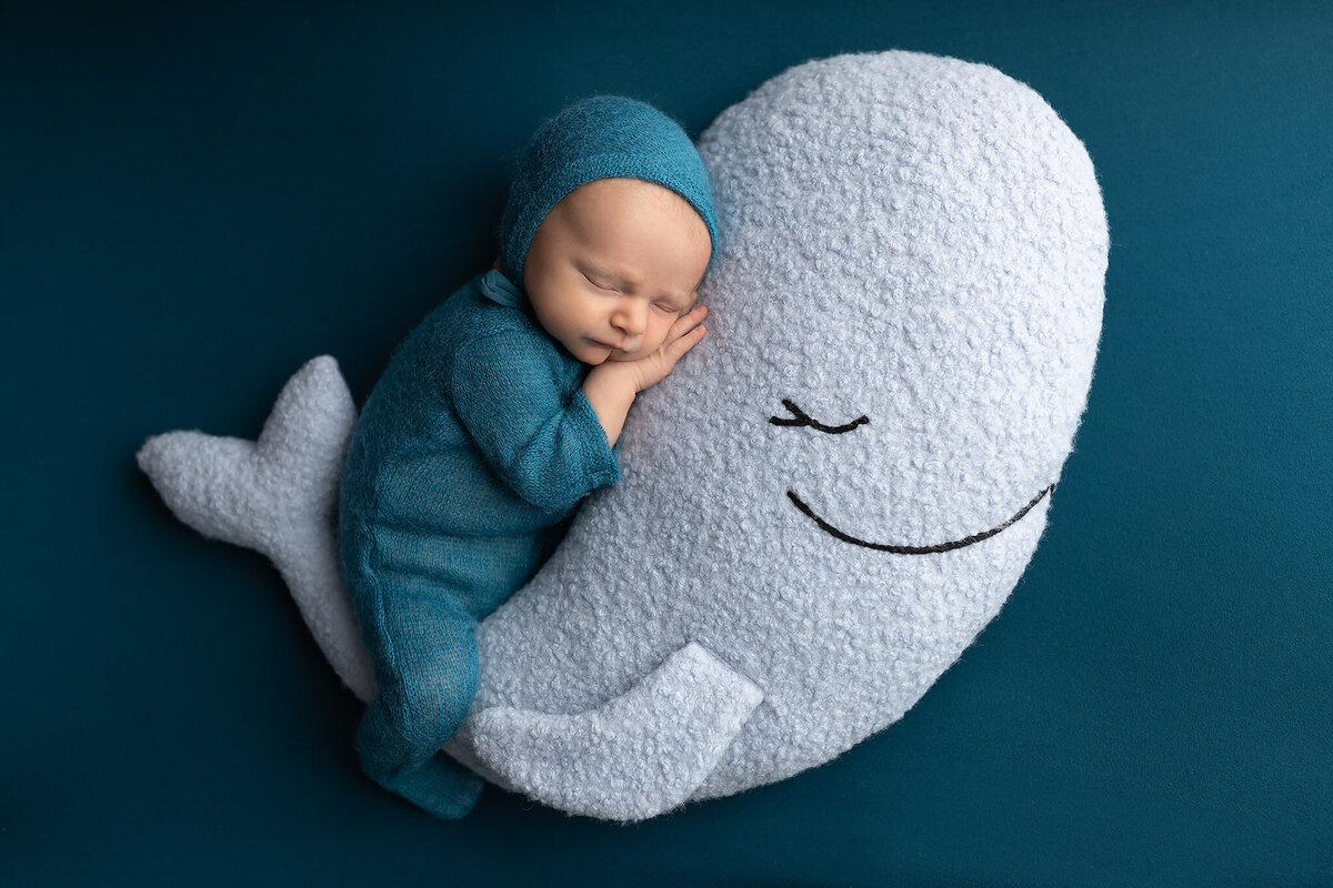 Peaceful newborn sleeping on a plush whale pillow in a cozy teal knit romper.