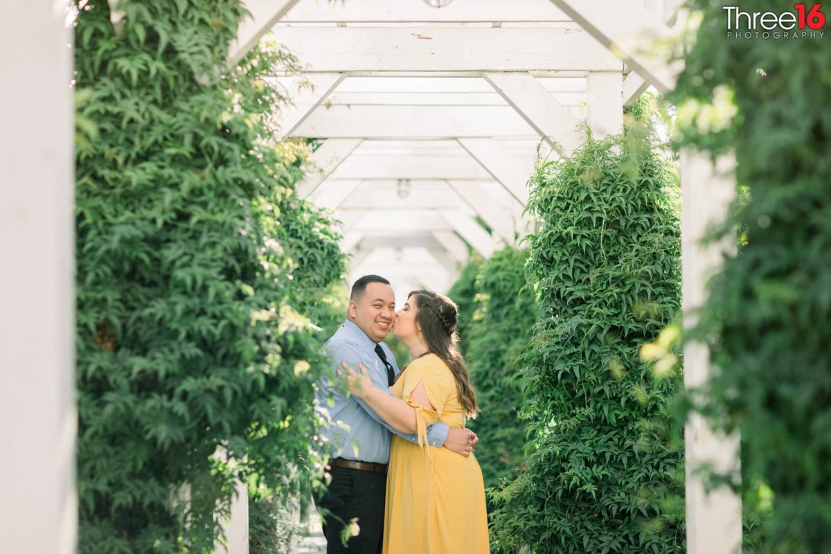 Bride to be kisses her fiance's cheek as he looks at the camera surrounded by green plants