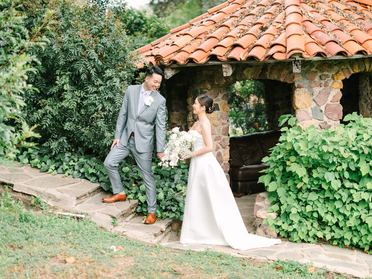 A joyful couple in wedding attire strolls hand in hand near a rustic stone gazebo with a red-tiled roof, surrounded by lush greenery, conveying romance and happiness.