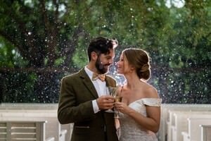 Professional St Augustine wedding photographers capture bride and groom toasting champagne in romantic rain shower with lush greenery backdrop