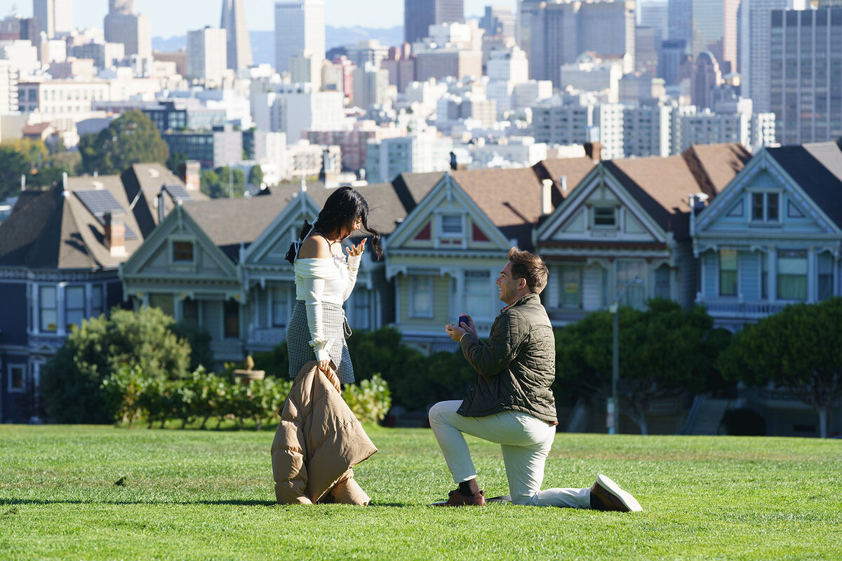 man on one knee proposing to a surprised woman with san francisco and the painted ladies in the background