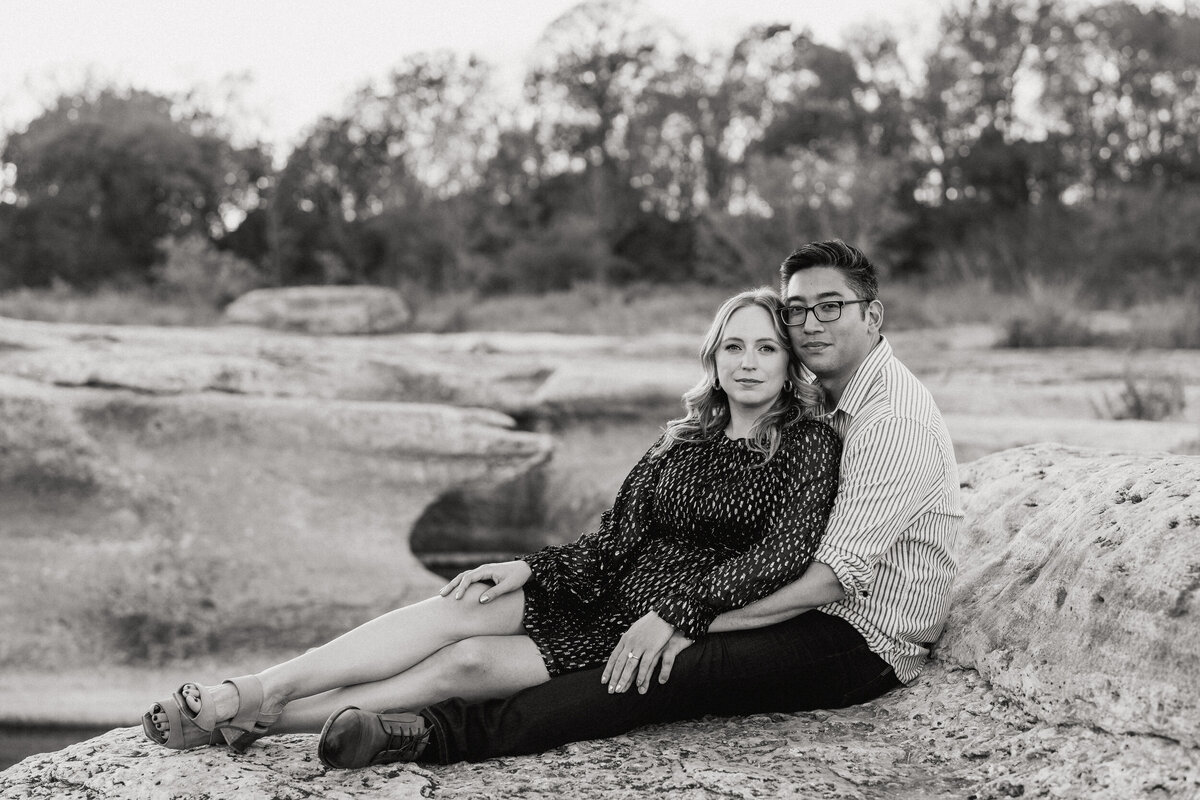 A black and white photograph of Rachel and Jason during their engagement session in McKinney Falls State Park in Austin, TX. The landscape made up of rock on the ground, carved out by the river below, which we can’t see. There are trees in the background. The couple is sitting in the ground with her sitting between his legs. She is leaning her back against him, their legs stretched out together, as they look towards the camera. Wedding and enagagement photography by Stacie McChesney/Vitae Weddings.