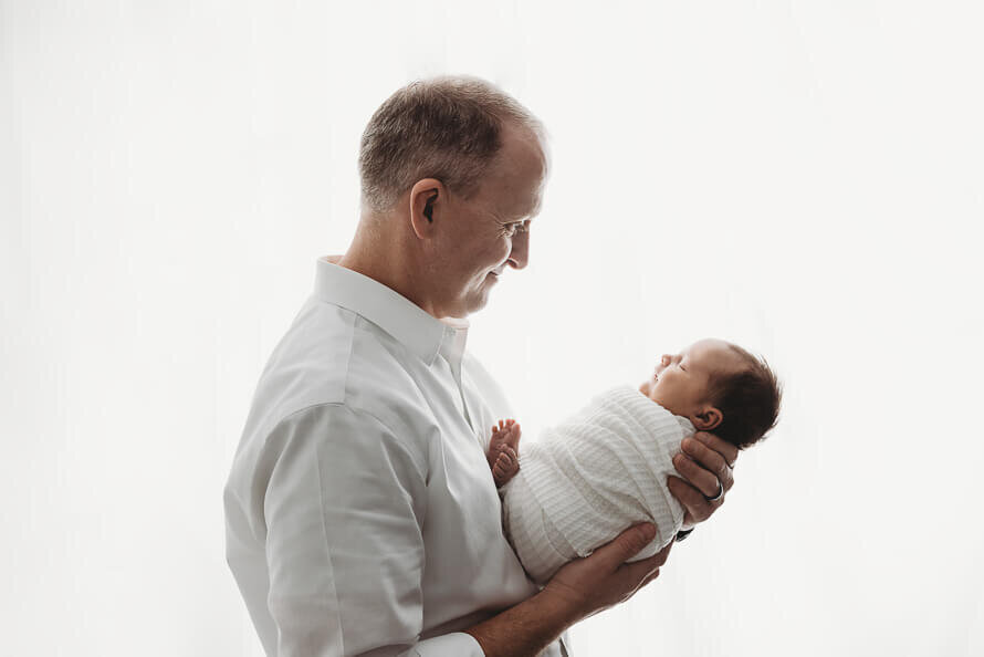 Dad looks at and holds his newborn baby in a backlit white studio.