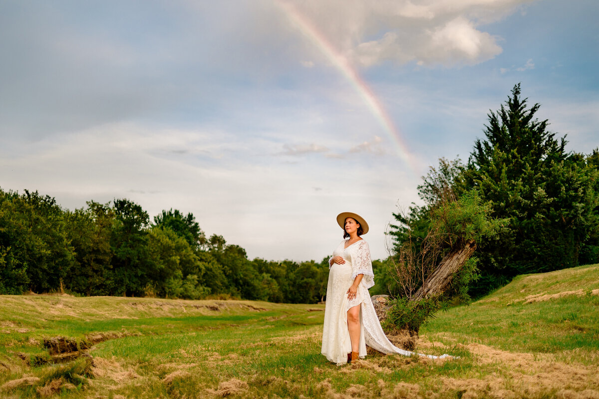 Photo of a pregnant woman in a forest with rainbow. The woman is wearing a long white dress and a brown hat