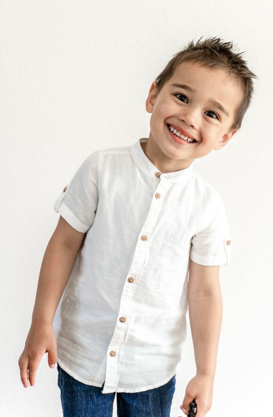 A close-up, high-angle portrait of a young boy with a wide, infectious smile. He is looking up at the camera with his head tilted to the side. He is wearing a white collared shirt with the sleeves rolled up and dark jeans, and the background is a simple white wall.