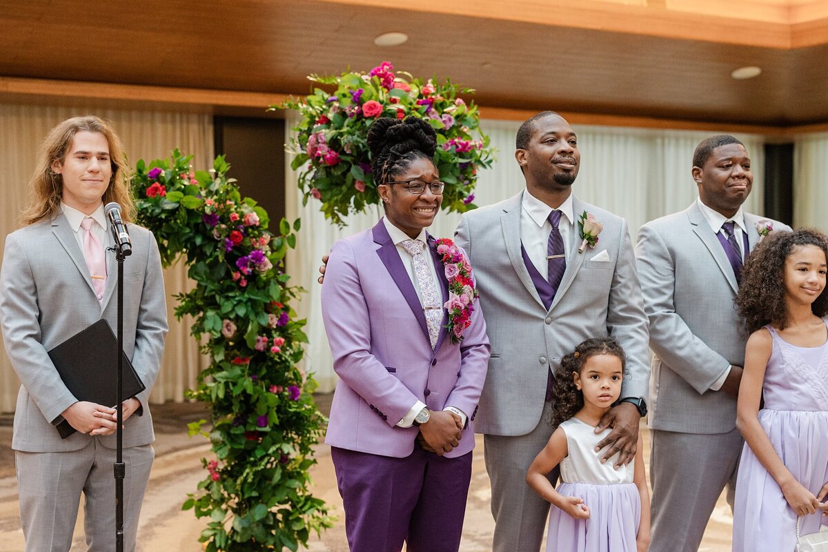 Candid shot of a bride reacting to seeing the other bride walk down the aisle during their wedding ceremony at a wedding in Dallas, Texas. The bride is overcome with emotion and is wearing a purple suit with a large, intricate boutonniere. Part of wedding part can be seen to her right alongside a few child attendants. The wedding officiant can be seen to her left. The whole group is backed by multiple, large floral arrangements.