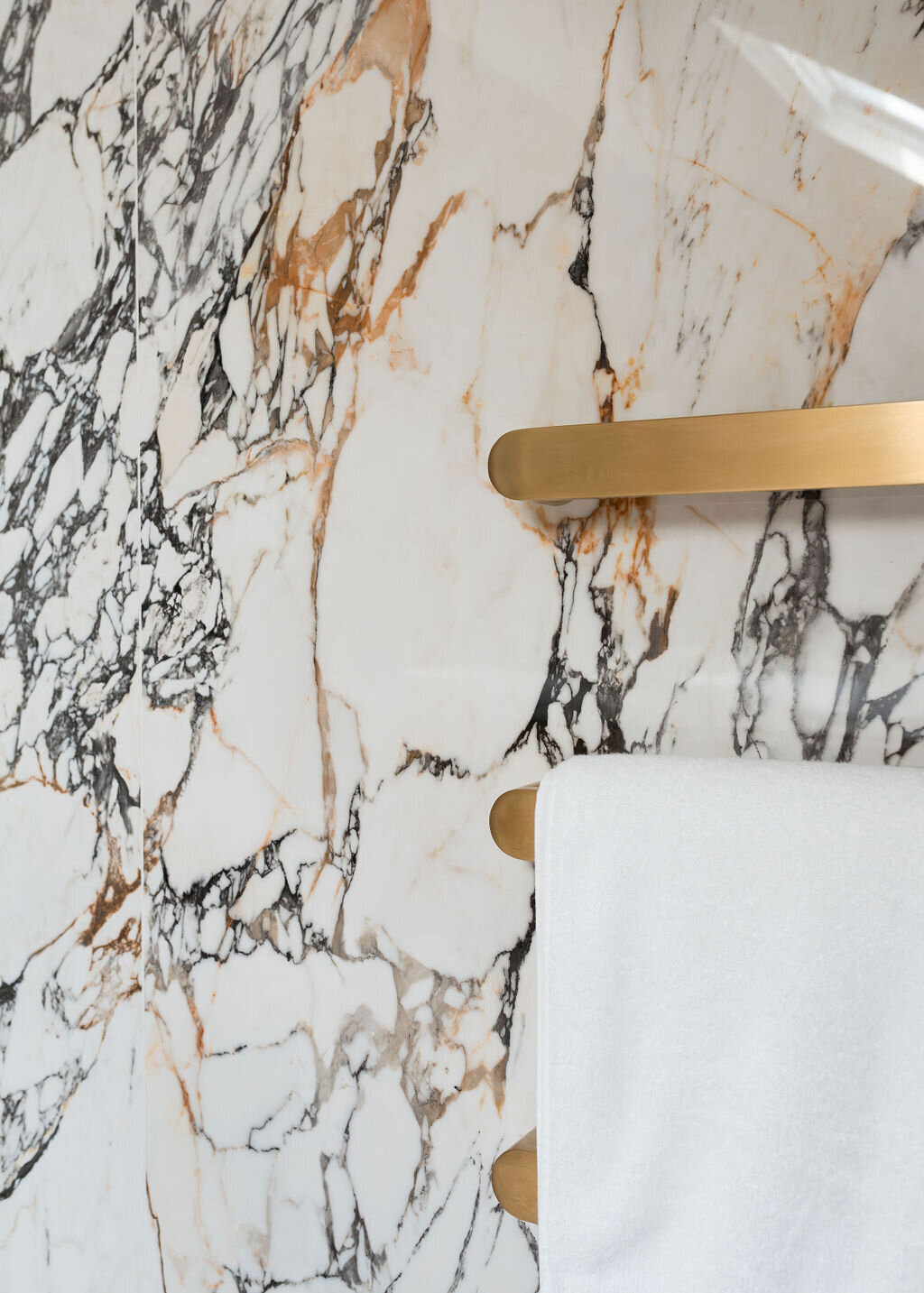 Modern gold towel bars are mounted on a white marble wall with black veining.