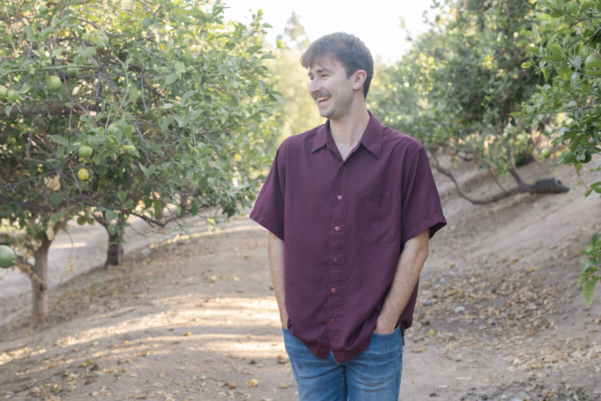 A young man wearing a maroon shirt and blue jeans standing on a dirt pathway between groves of lime trees with his hands in his pockets, laughing as he's looking away from the camera.
