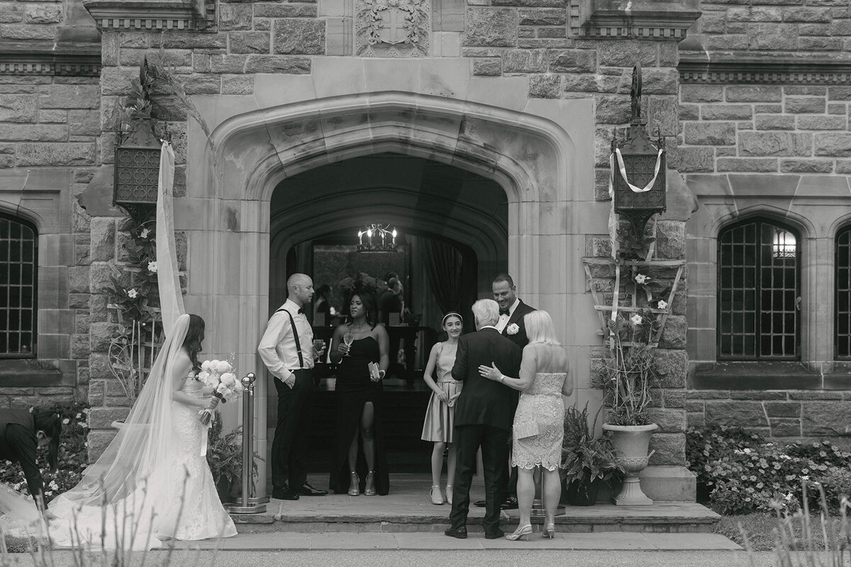 Elegant documentary-style wedding moment of guests gathering beneath the grand stone entrance of Mill Neck Manor in Oyster Bay, New York