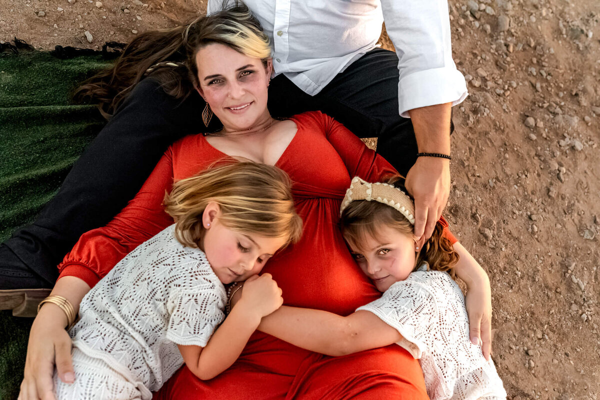 Daughters lying with their mom on blanket in the Arizona desert, hugging her baby bump