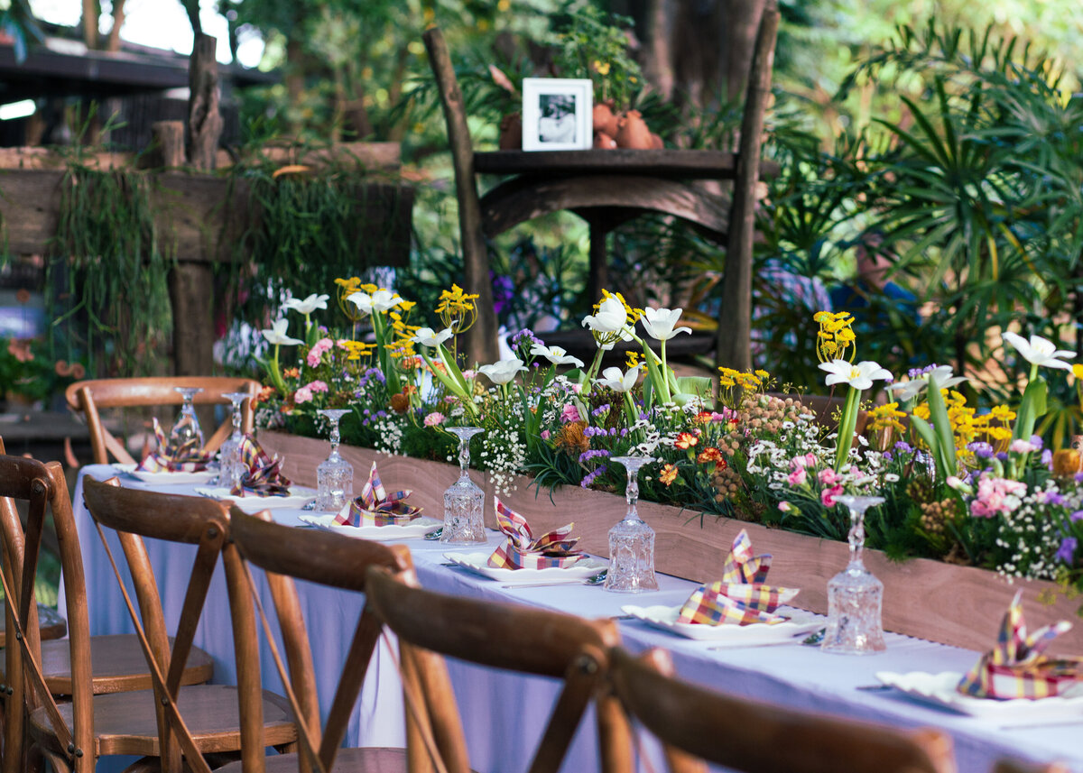 An event table with a blue tablecloth and colourful wild flowers in a central wooden planter for an outdoor wedding or party.