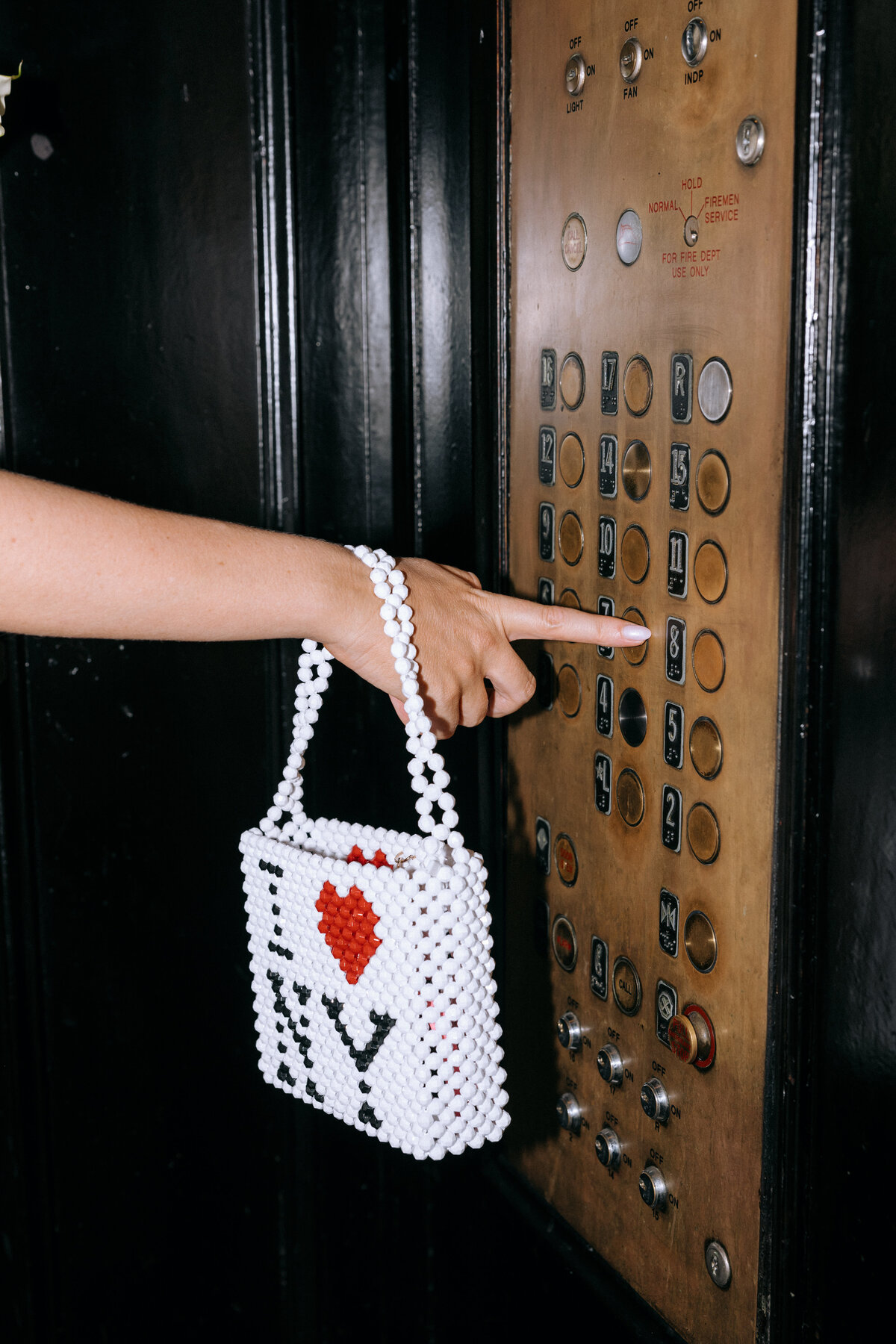 Bride holding an I Love NY beaded bag while pressing a button in a vintage elevator on her New York City wedding day

