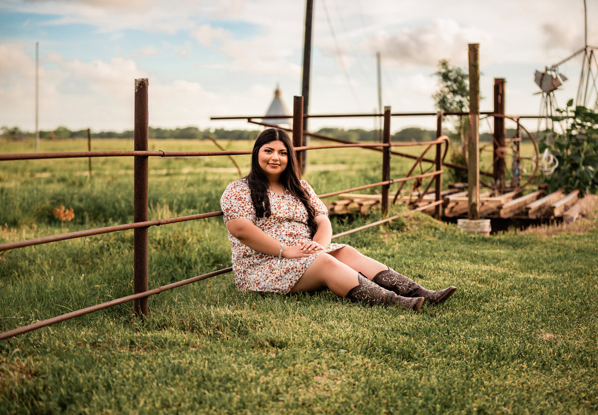 A Houston senior sits leaning against a metal fence post with a cloudy blue sky behind her.