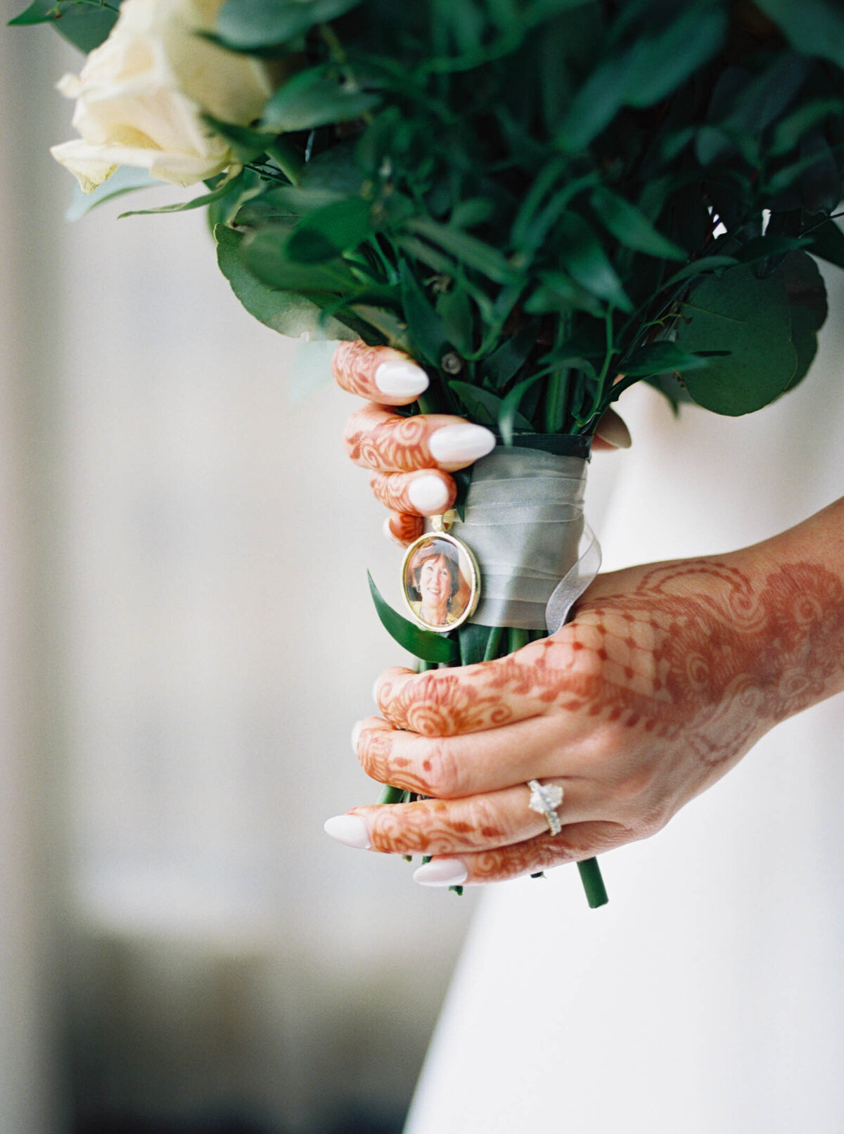 Film image of a bride's mehndi covered hands holding a bouquet with a pendant that memorializes her deceased mother.