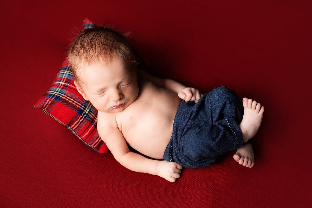 Newborn boy in blue jeans posed on a red blanket with matching plaid pillow