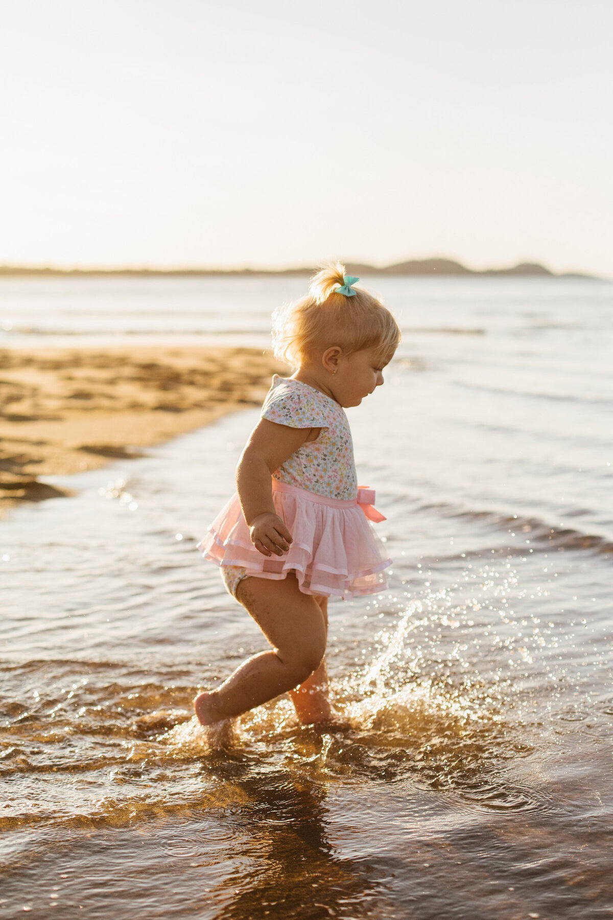 Little girl in a pink tutu joyfully running along the sandy beach with sunlight behind her.
