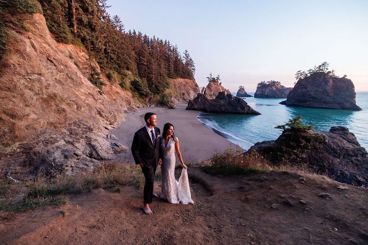 A bride and groom stand on a cliff side overlooking the sunset. 
