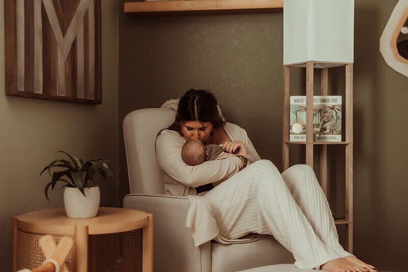 Mom kissing newborn baby boy in rocking chair in nursery