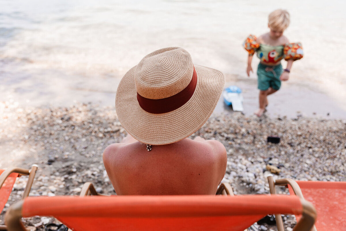 Moeder met hoed zit in oranje stoel aan het meer en kijkt toe hoe jongetje met zwembandjes in het meer speelt