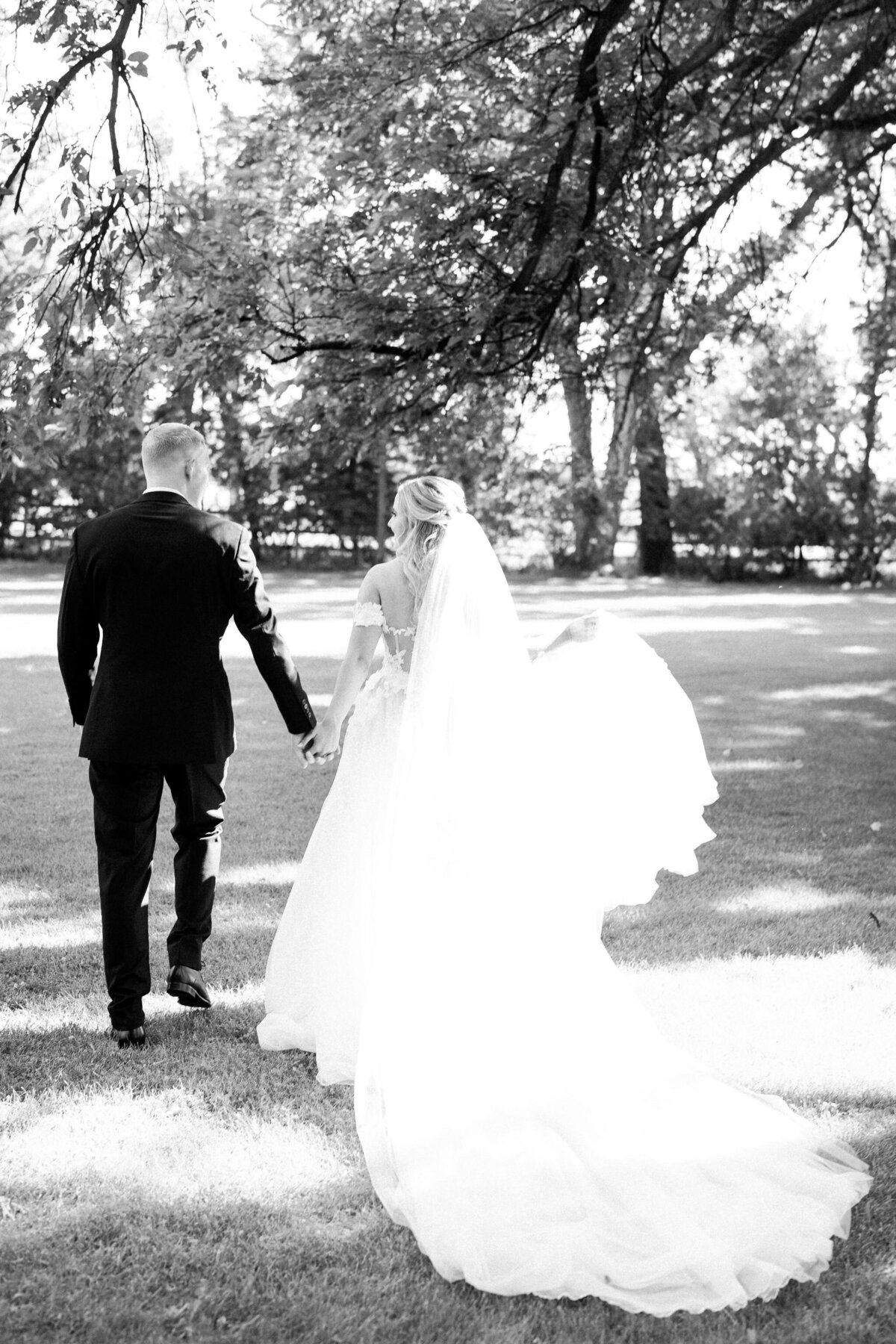 A bride and groom hold hands while walking on a grassy area under trees at their Norland Estate wedding in Calgary. The bride's long veil and train flow behind her. The image is in black and white.