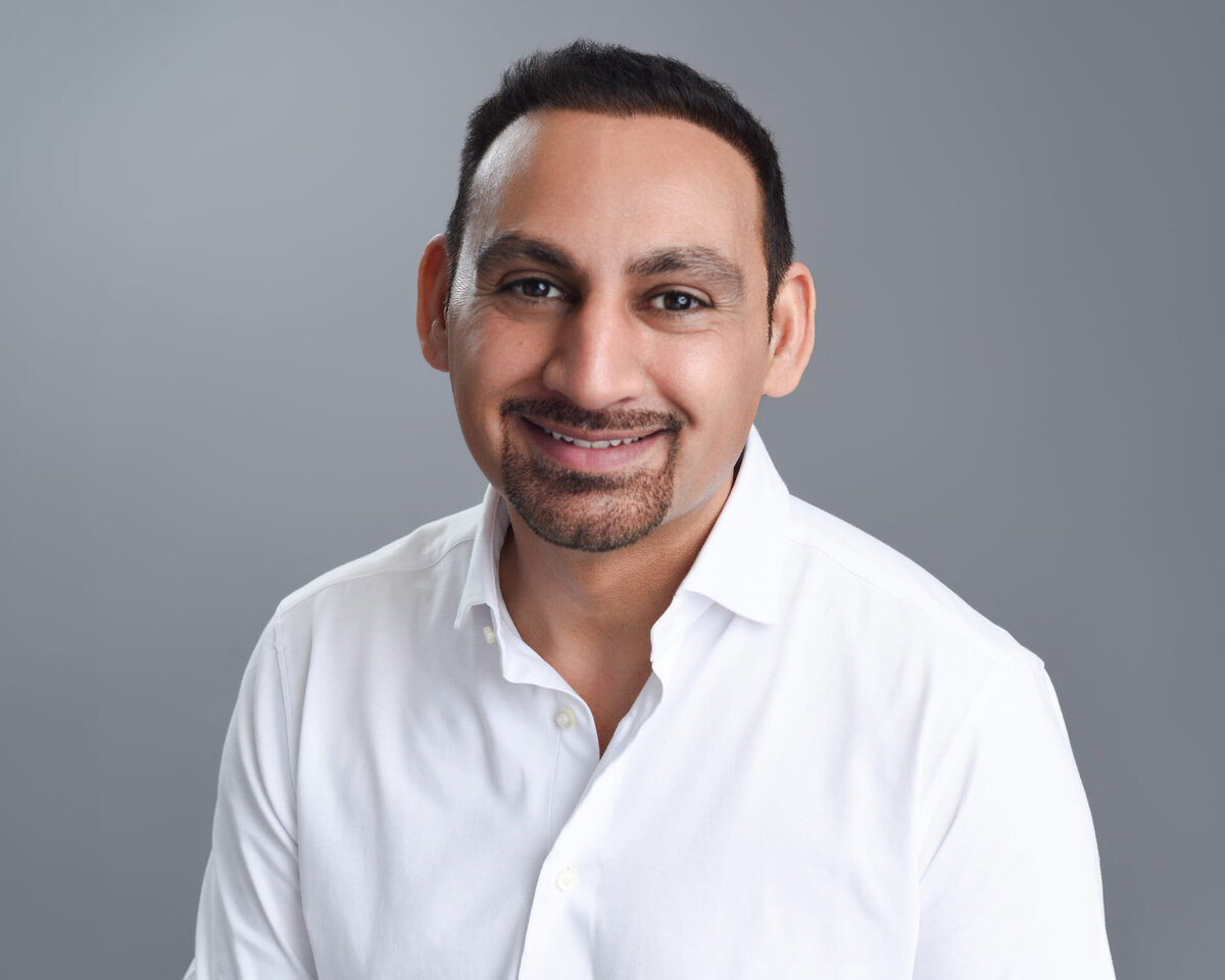 Professional headshot of an individual wearing a crisp white button-up shirt, posed against a neutral grey backdrop. The subject’s direct gaze and polished styling create an approachable yet confident presence—ideal for business branding and corporate photography