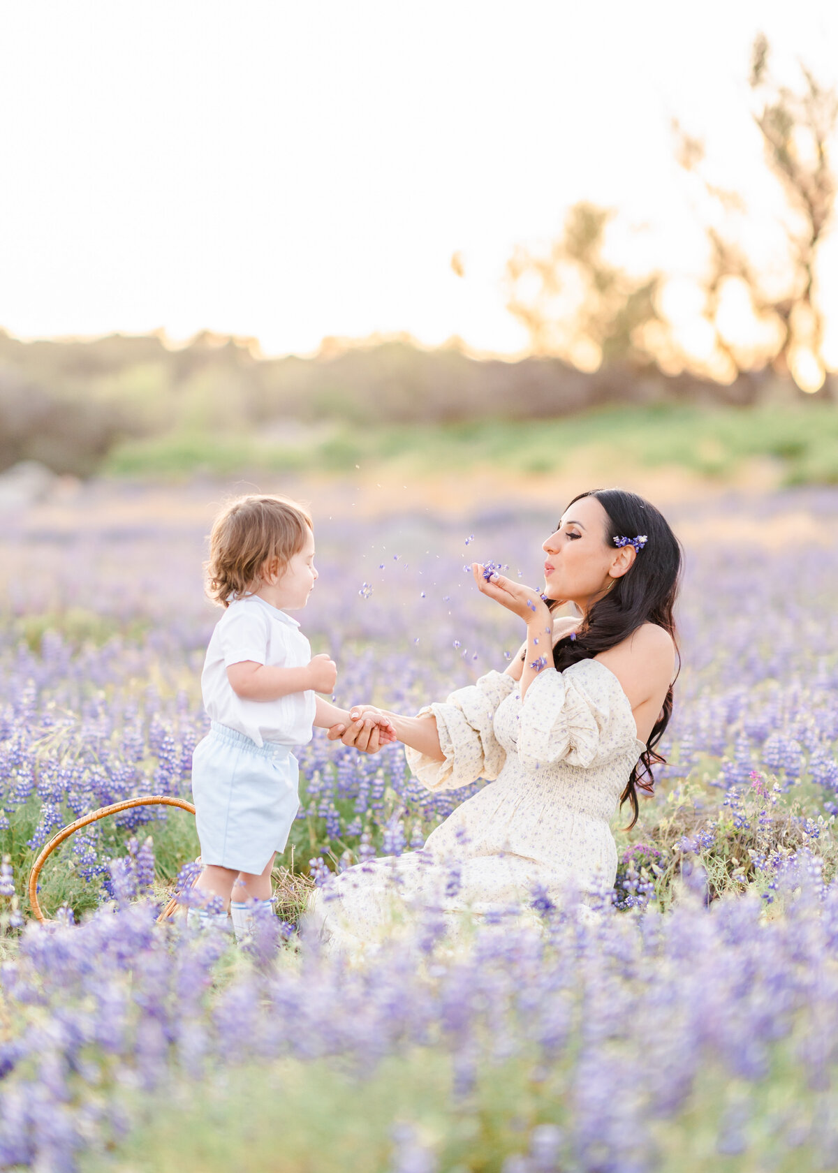 A mother kneels in a field of purple lupines blowing flower petals at her son who is standing holding her hand photographed by bay area photographer, Light Livin Photography.