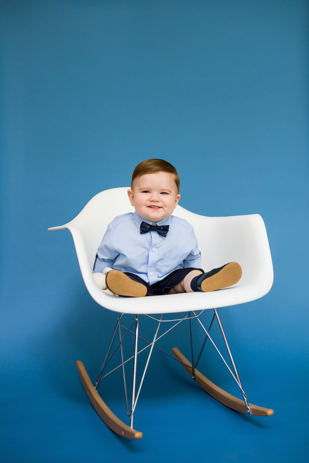 Baby sitting in Eames Molded Plastic Armchair Rocker chair on blue backdrop for baby's first year photography session.